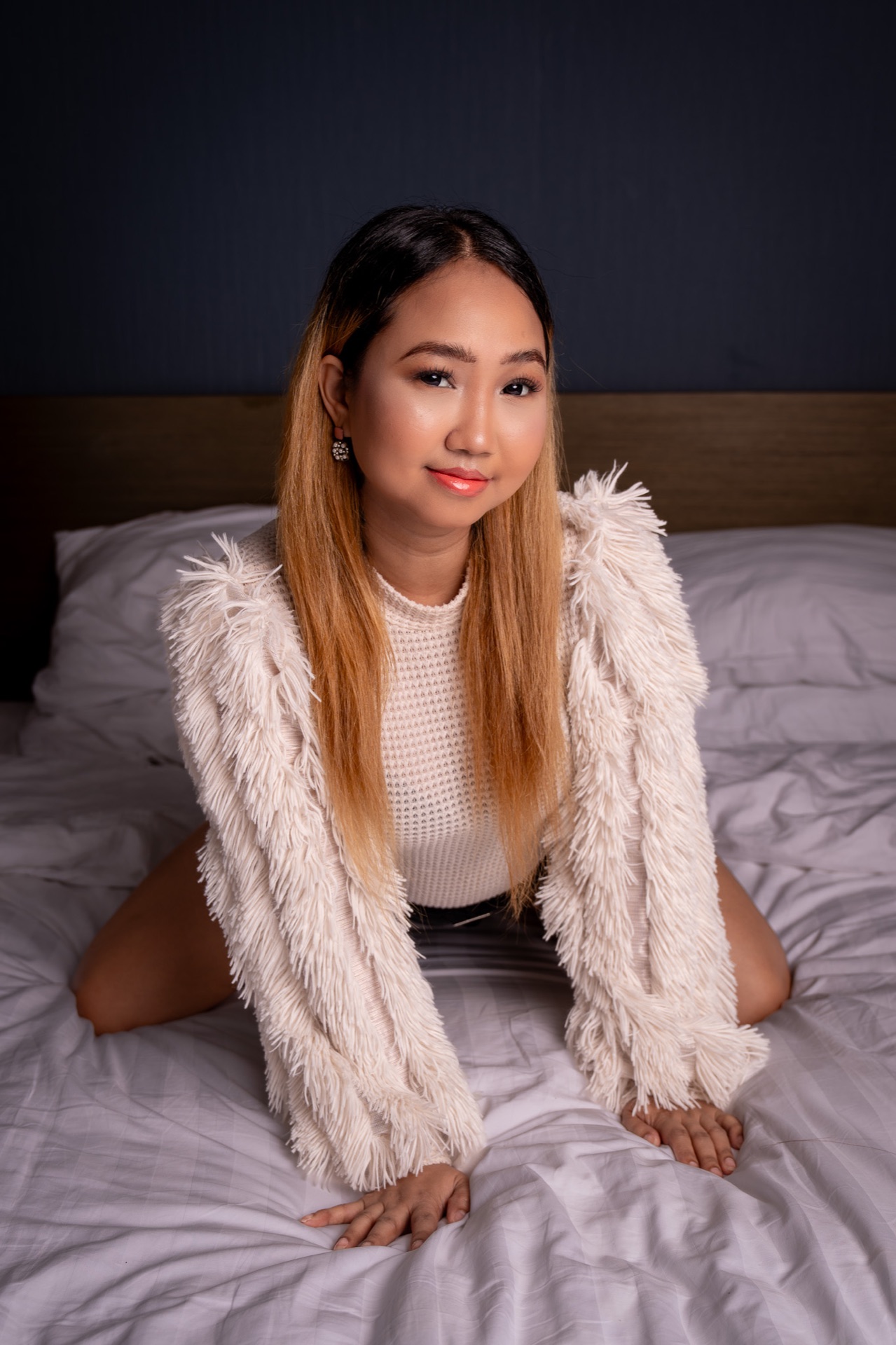 Lovely sits on a bed, smiling toward the camera, wearing a white textured sweater and fringed jacket, with long highlighted hair.