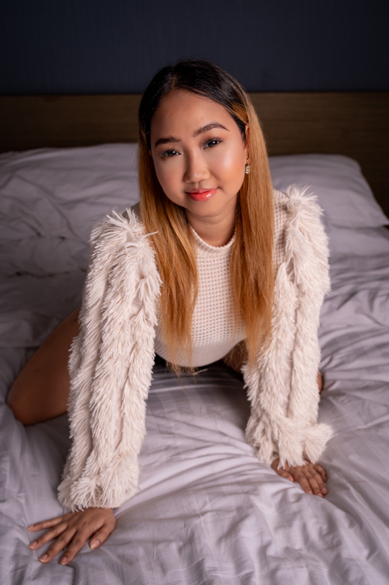 Lovely seated on a bed, facing the camera with a slight smile, wearing a white textured sweater and fringed jacket.