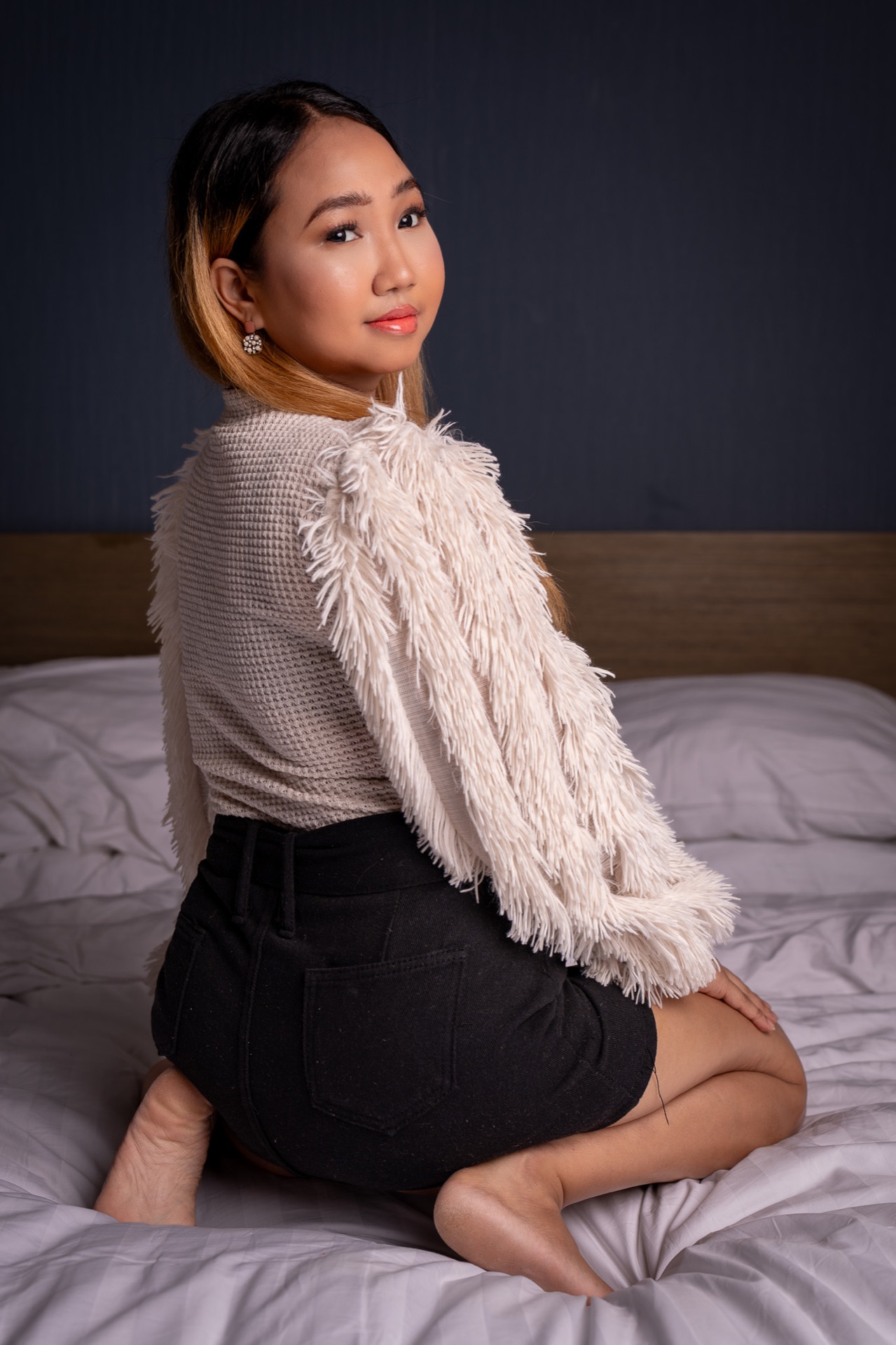 Lovely sits sideways on a bed, looking toward the viewer, wearing a textured sweater with fringed sleeves and a black skirt.