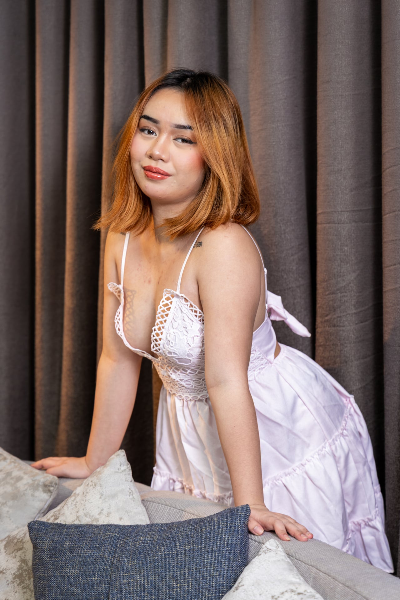 Kath poses beside a sofa in a white lace-trimmed dress, looking toward the camera against dark curtains in Makati.