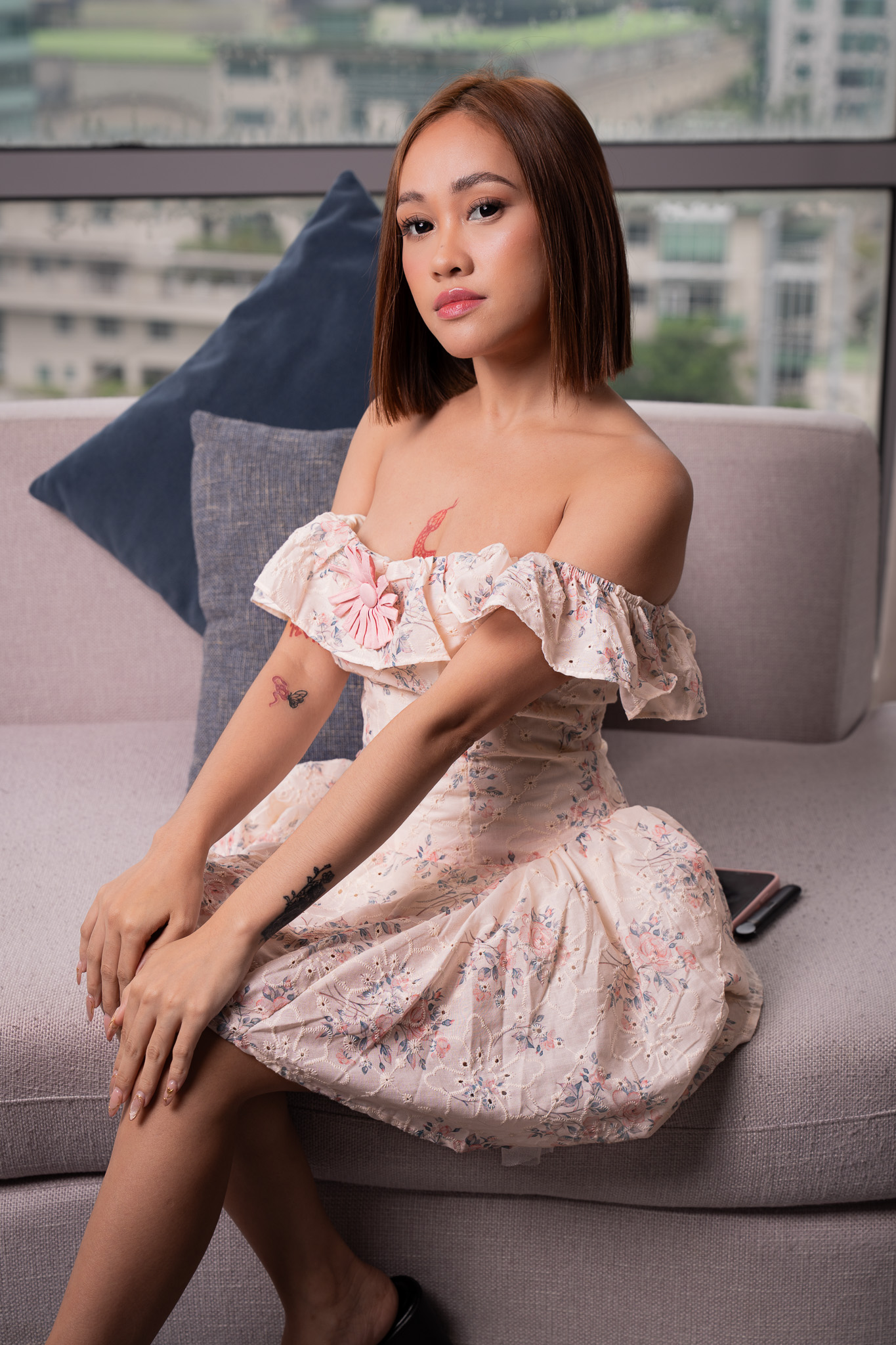 Whisky sits on a gray sofa in a floral off-shoulder dress, gazing toward the camera with city buildings visible through the window.