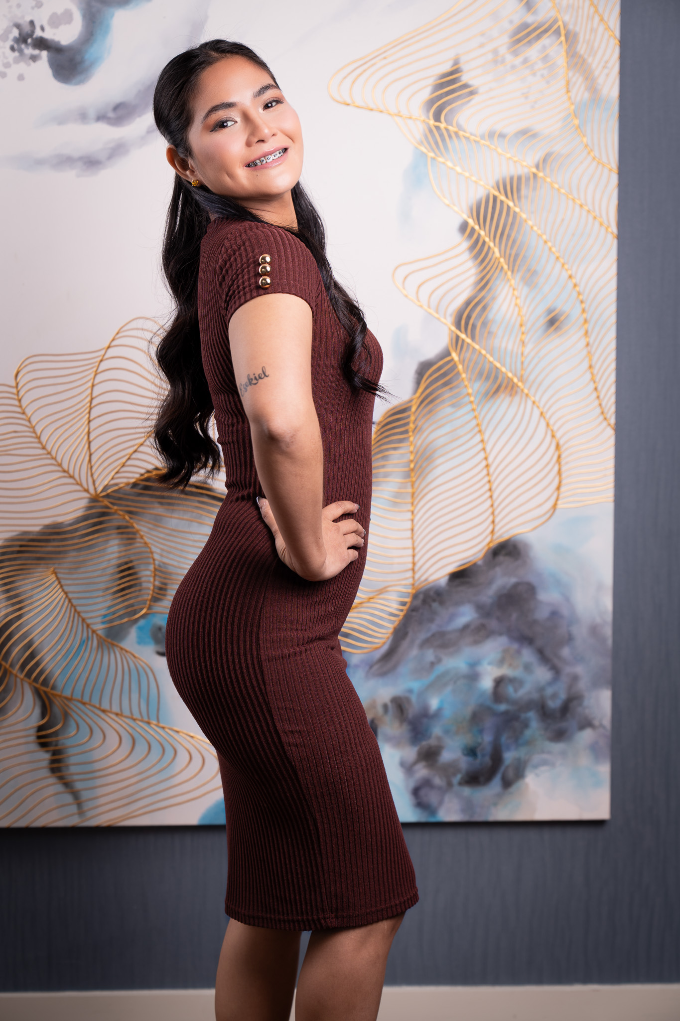 Yana stands in three-quarter profile, smiling in a ribbed burgundy dress before abstract gold-line wall art.